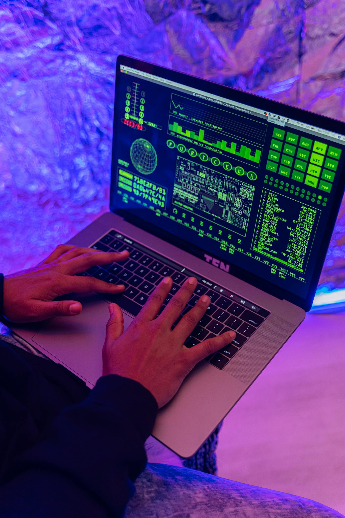 Close-up of hands typing on a laptop displaying green code, indoors, purple lighting.
